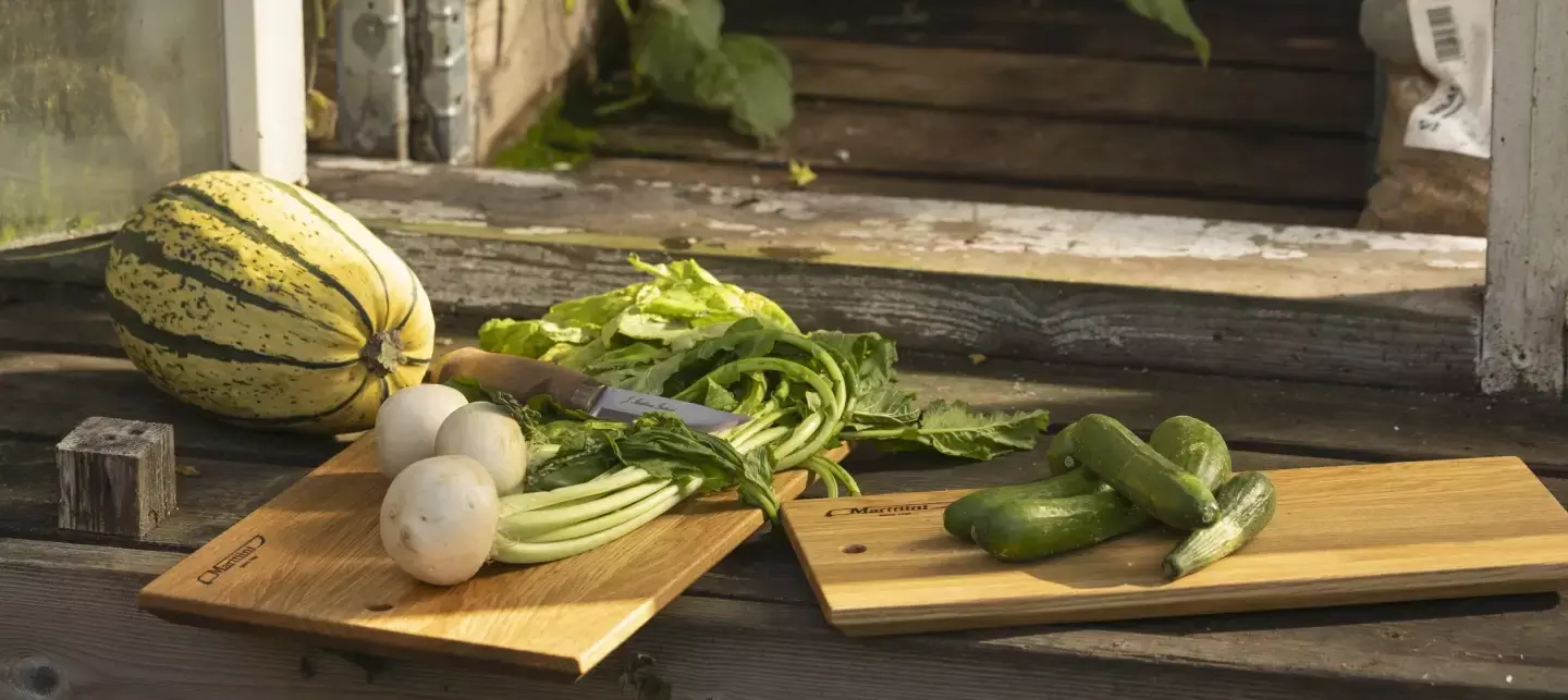 Planche à découper en chêne Marttiini appuyée contre un encadrement de fenêtre rustique, à côté d’un bol en céramique rempli d’herbes vertes fraîches dans un cadre de jardin.