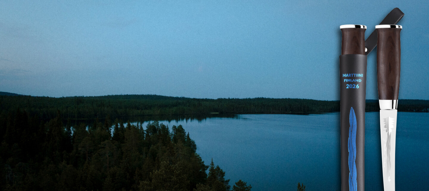 Marttiini Järvi Vuosipuukko 2026, jossa tummapuinen kahva ja musta tuppi sinisellä aaltokuviolla, on aseteltu järvimaisemataustalle.