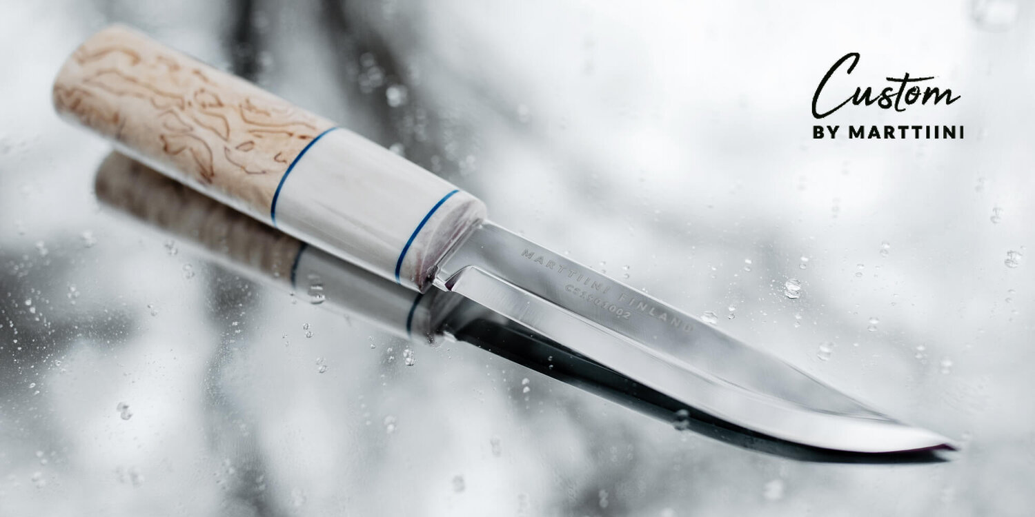 Gros plan sur un couteau Marttiini Custom Shop avec manche clair et lame polie se reflétant sur une surface mouillée.