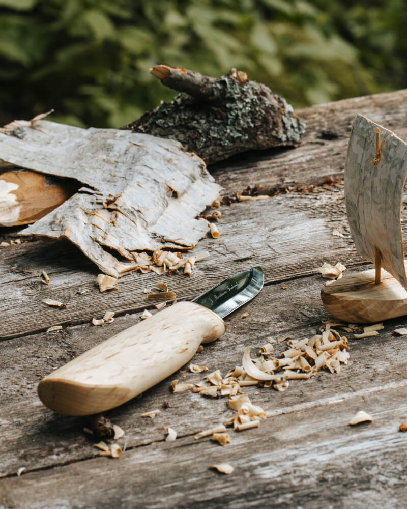 La photo montre le couteau pratique de Marttiini posé sur une table en bois. Avec ce couteau, il vient de sculpter un bateau en écorce de bouleau, dont on voit des copeaux sur la table.