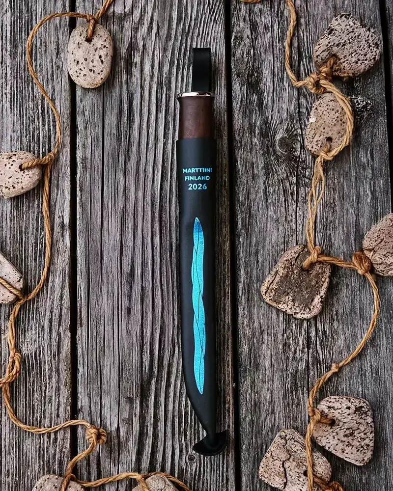 Marttiini Finland 2026 Messer in schwarzer Scheide mit blauem Ziermuster, auf einer verwitterten Holzoberfläche neben einer Steinschnur platziert.