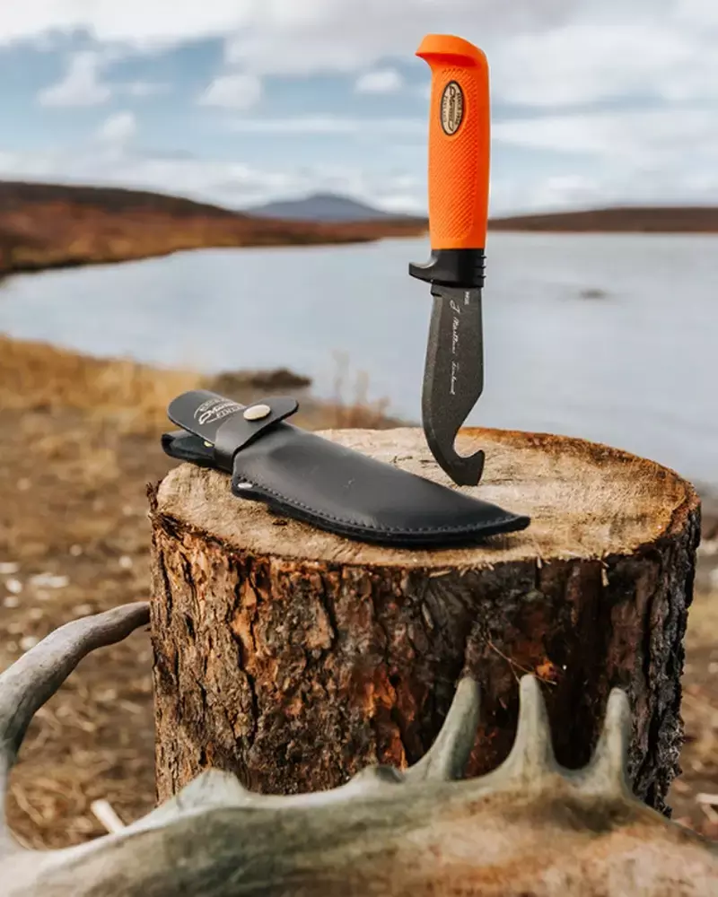 Marttiini Jagdmesser mit orangefarbenem Griff und schwarzer Klinge, auf einem Baumstumpf in einer Wildnislandschaft am See platziert.