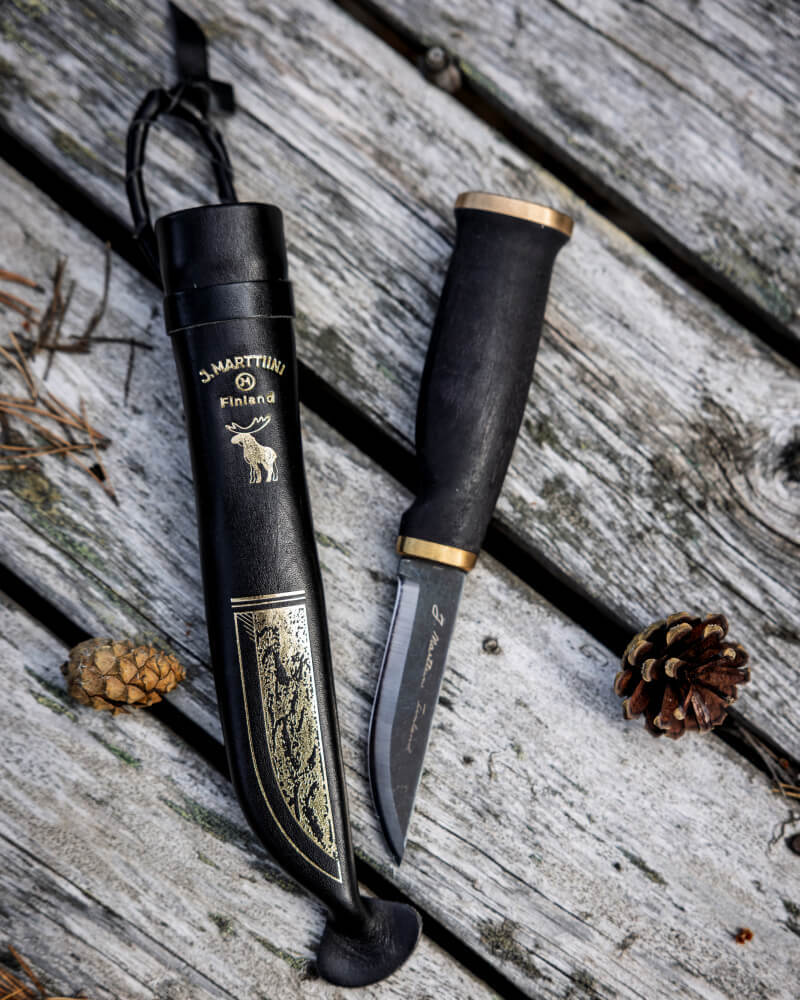 A Marttiini Moose Black knife with a dark handle and brass details lying next to its black leather sheath with gold-embossed decoration on a weathered wooden surface outdoors.