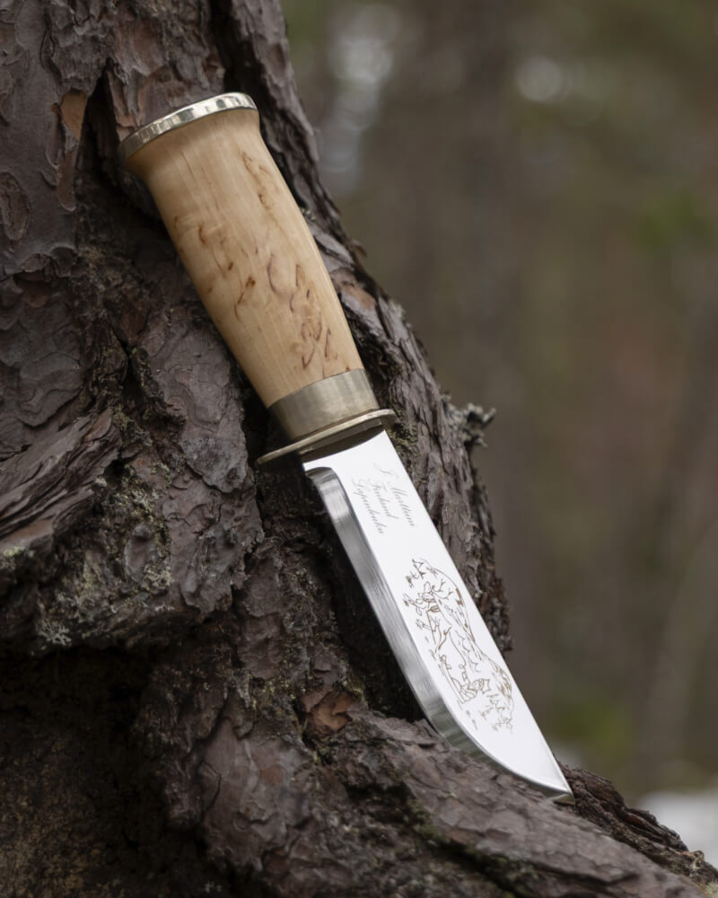 Ein Marttiini-Messer mit einem Griff aus Maserbirke und einer gravierten Klinge, das im Freien in der Rinde einer Kiefer ruht.