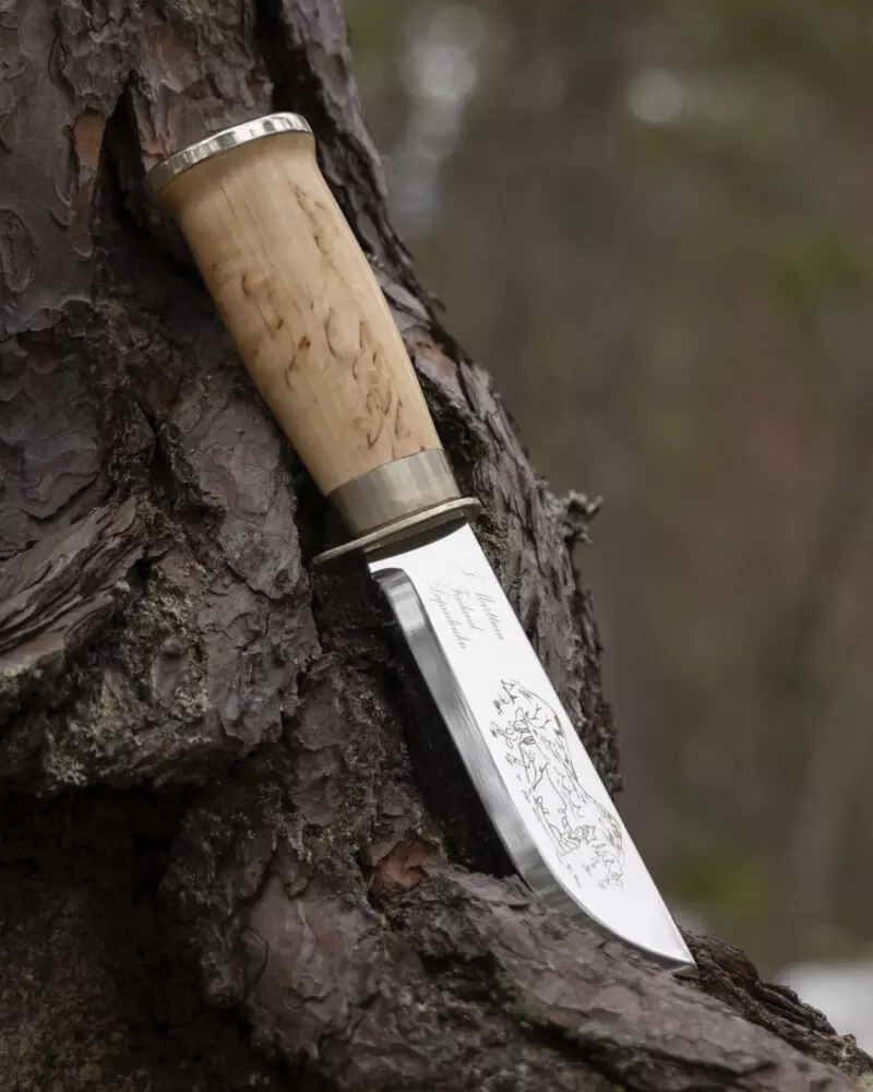 Marttiini Messer mit gravierter Klinge und Maserbirken-Griff, an einen Baumstamm gelehnt.